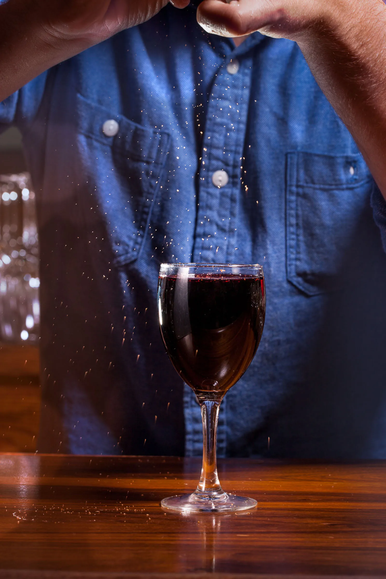 Bartender in a denim shirt crumbles garnish over a dark red drink, amber particles frozen mid-air.