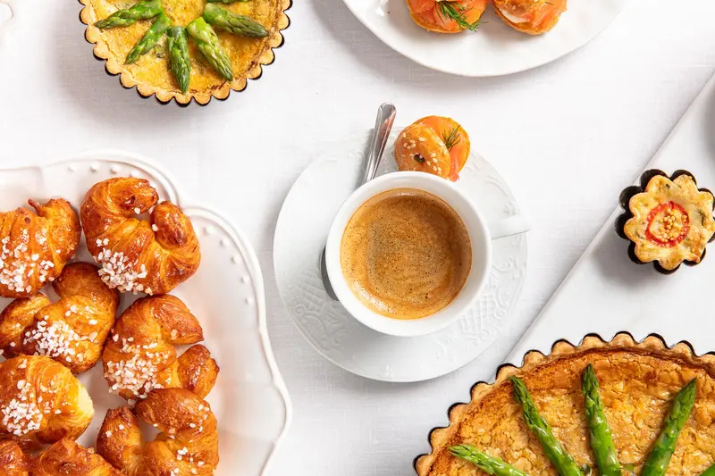 Breakfast spread shot from above: pastries, asparagus tarts, and an espresso on a white table.