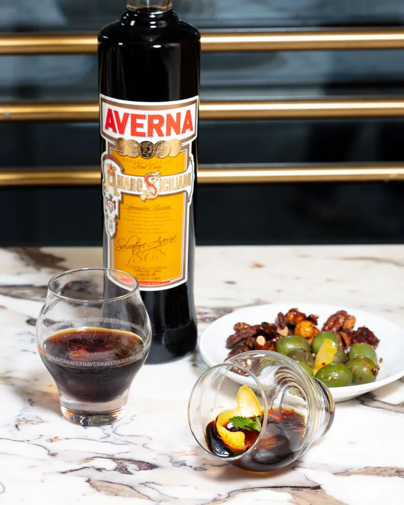 Averna amaro bottle with its yellow label in sharp focus, a poured amaro and a tipped glass with orange peel on marble.