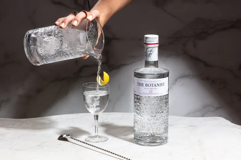 Gin poured from a crystal mixing glass into a martini glass beside The Botanist gin bottle on marble