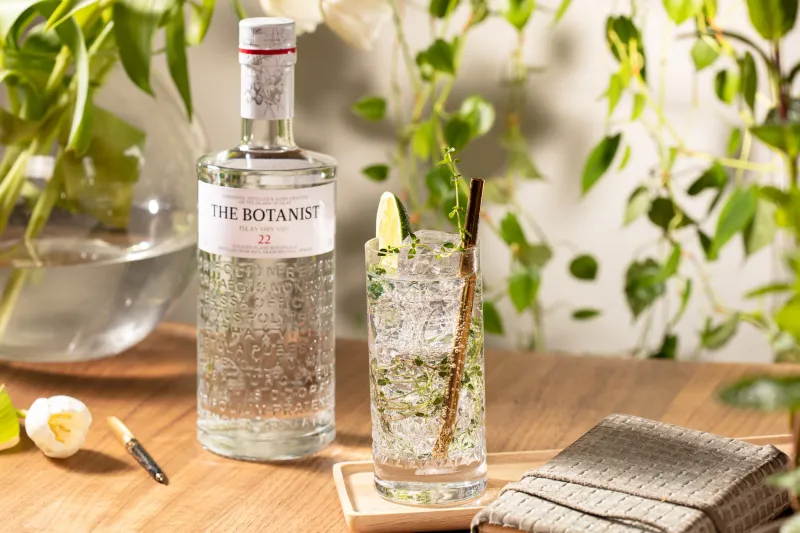 The Botanist gin bottle and a tall iced gin cocktail on a wooden table, trailing plants behind