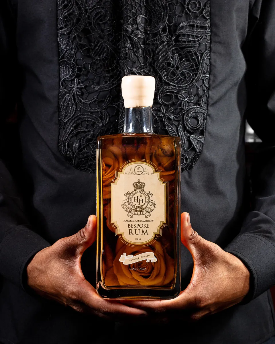 Hands holding a bottle of HH Bespoke rum against a dark embroidered jacket.