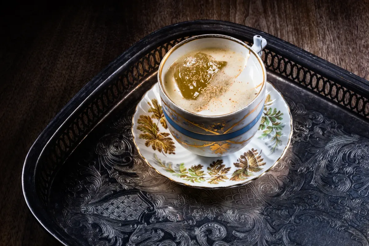 A cocktail in a porcelain teacup on an engraved silver tray, topped with grated nutmeg and a large clear ice piece.