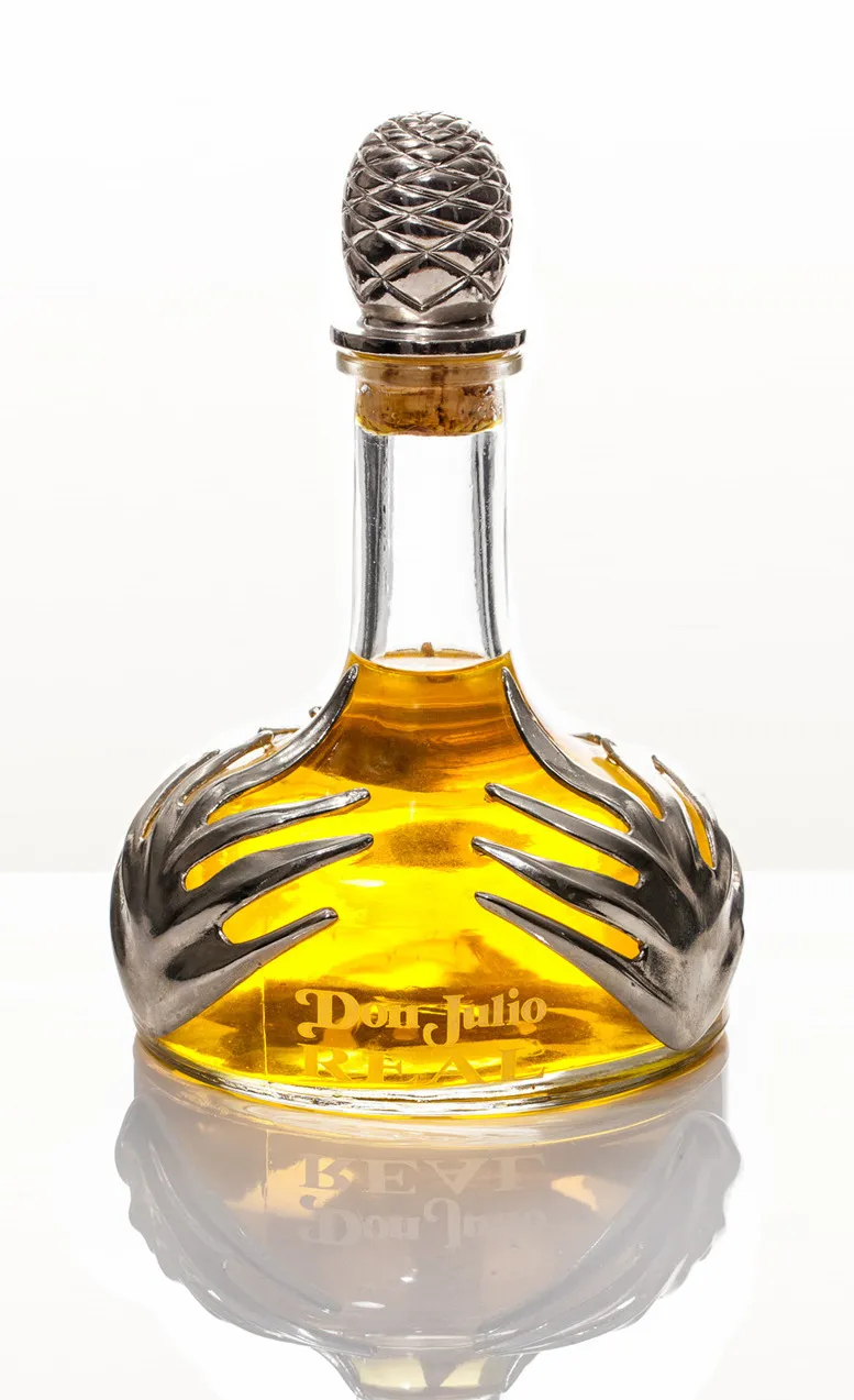 Don Julio Real tequila decanter on white, its silver hand-shaped collar and amber liquid mirrored in the floor reflection.
