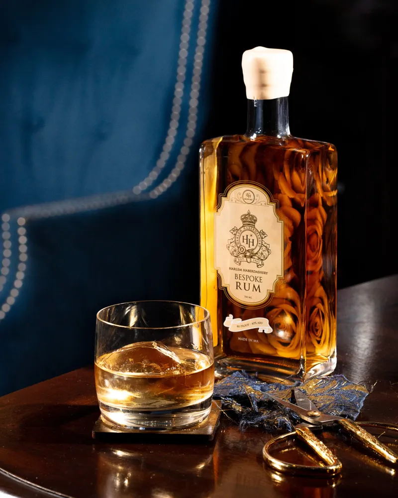A bottle of HH Bespoke rum beside a rocks glass with a single large ice cube, blue velvet banquette behind.