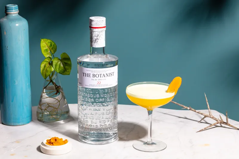 The Botanist gin bottle beside a yellow cocktail with an orange peel garnish against a teal wall