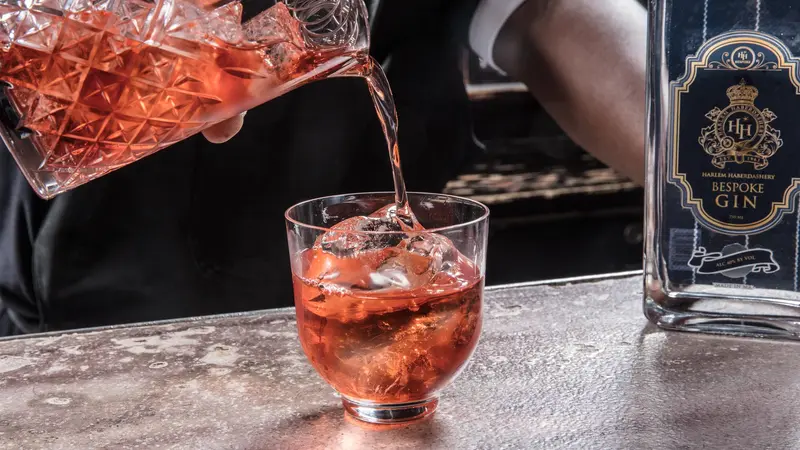 Negroni poured from a cut-glass mixing glass over ice beside a bottle of HH Bespoke gin.