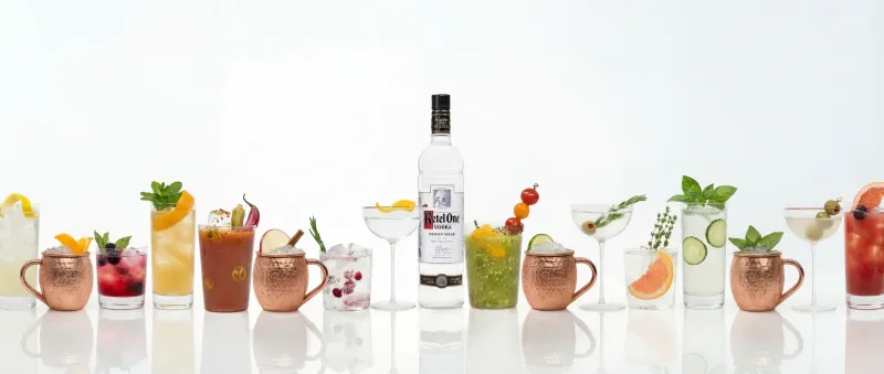 Ketel One vodka bottle at the center of a lineup of cocktails and copper mugs on white.