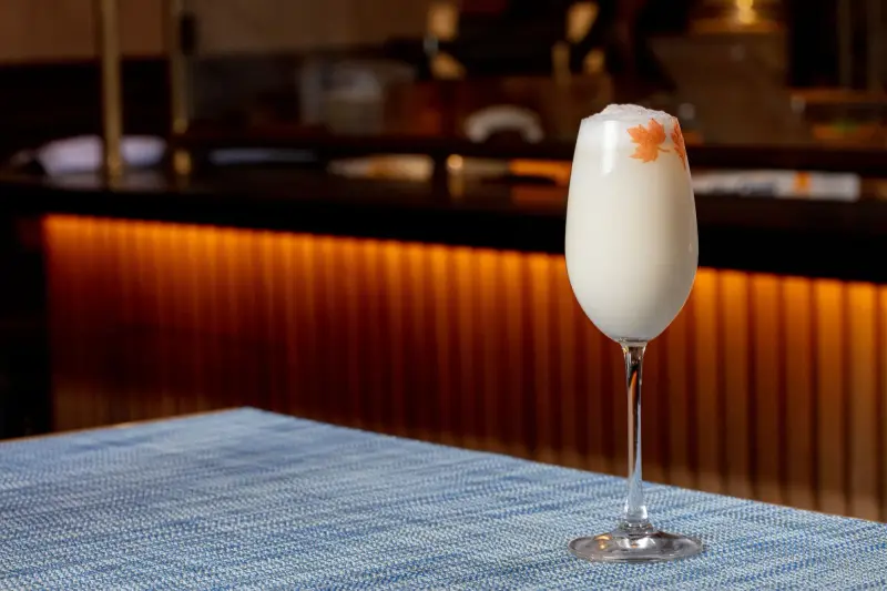 White frothy cocktail with an orange leaf stencil in a tall stemmed glass on a blue woven placemat, a warmly lit bar behind