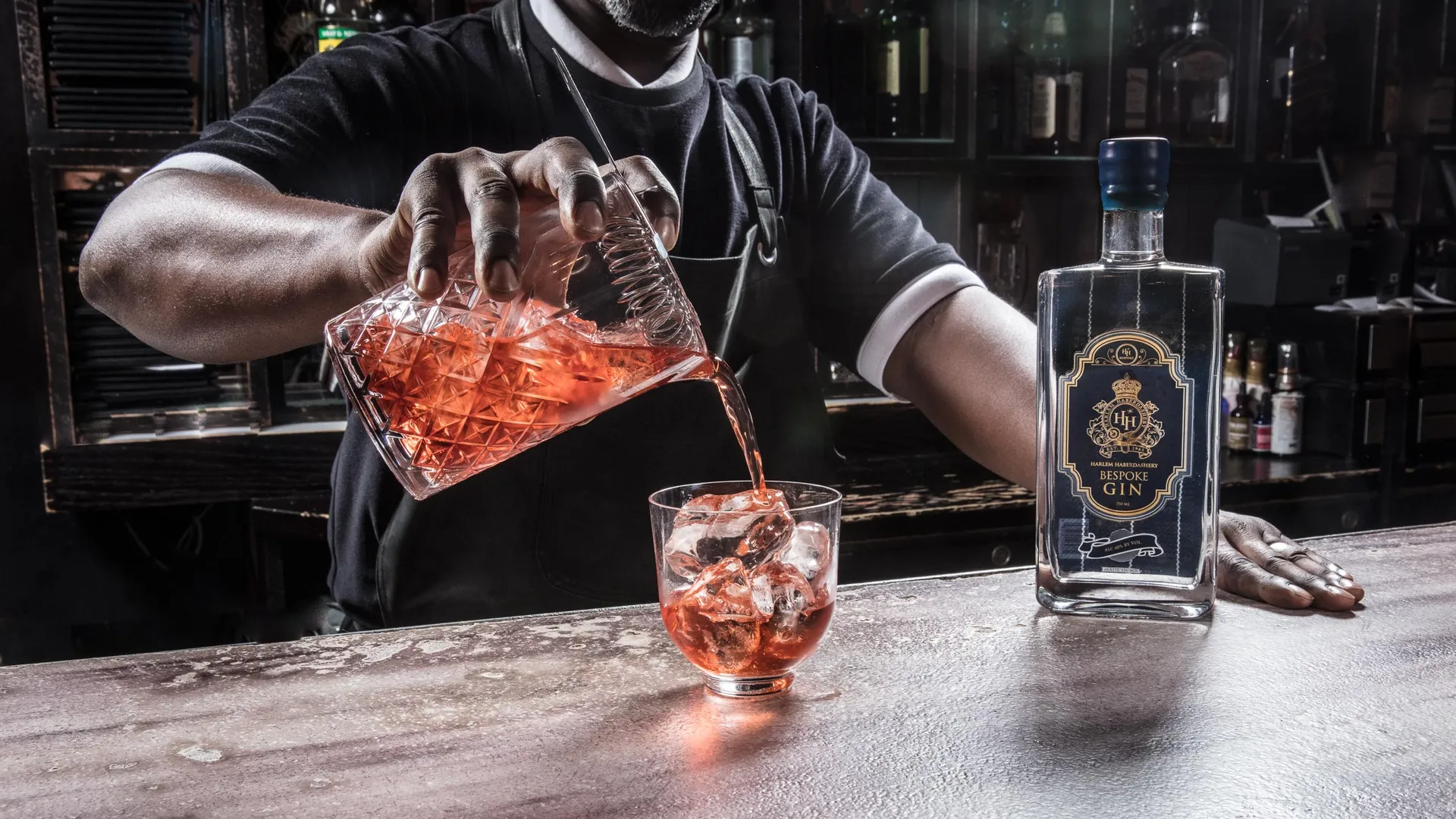 A bartender pours a negroni from a cut-crystal mixing glass over ice, a bottle of HH Bespoke gin on the bar.