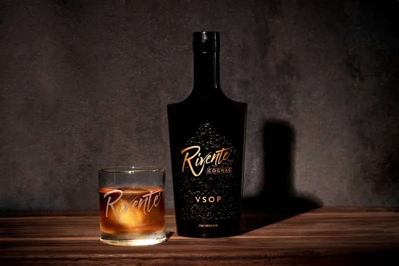 Rivente VSOP cognac bottle beside a pour over a large ice cube in a rocks glass, on dark wood