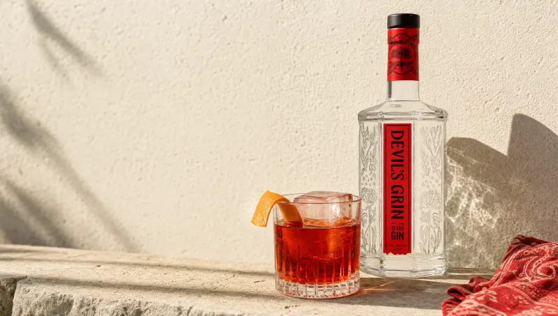 Generated frame for Devil's Grin gin: a negroni with an orange twist beside the bottle against a sunlit limestone wall.