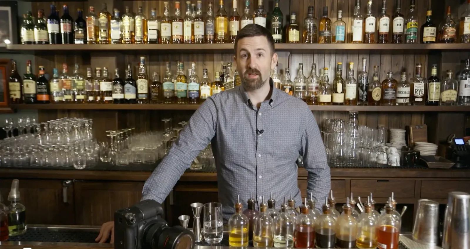 Brent Herrig behind a cocktail bar with his camera, bitters bottles lined up in front of him.