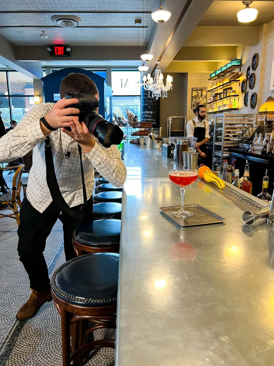 Brent Herrig photographs a red cocktail in a coupe on a zinc bar while a bartender works across the bar.