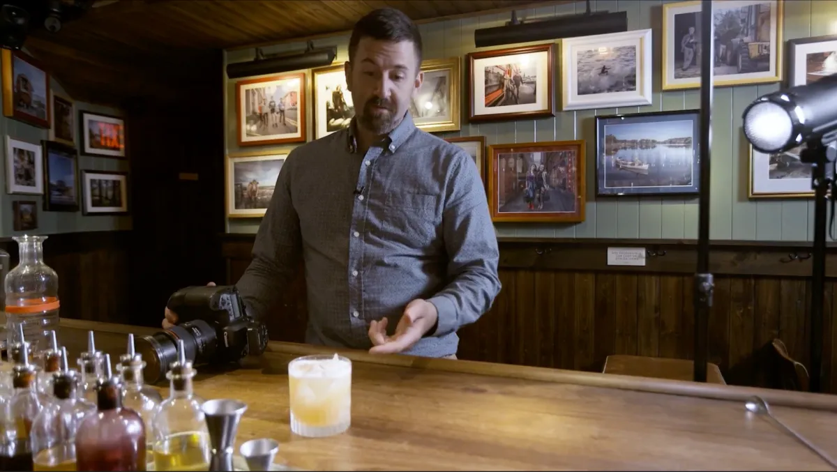 Brent Herrig explains commercial lighting at a wooden bar, camera in hand, strobe and rocks glass beside him.