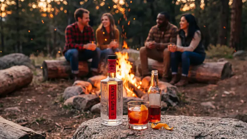 AI campaign frame for Devil's Grin gin: friends talk around a campfire at dusk, the bottle and a negroni on a granite rock in front.
