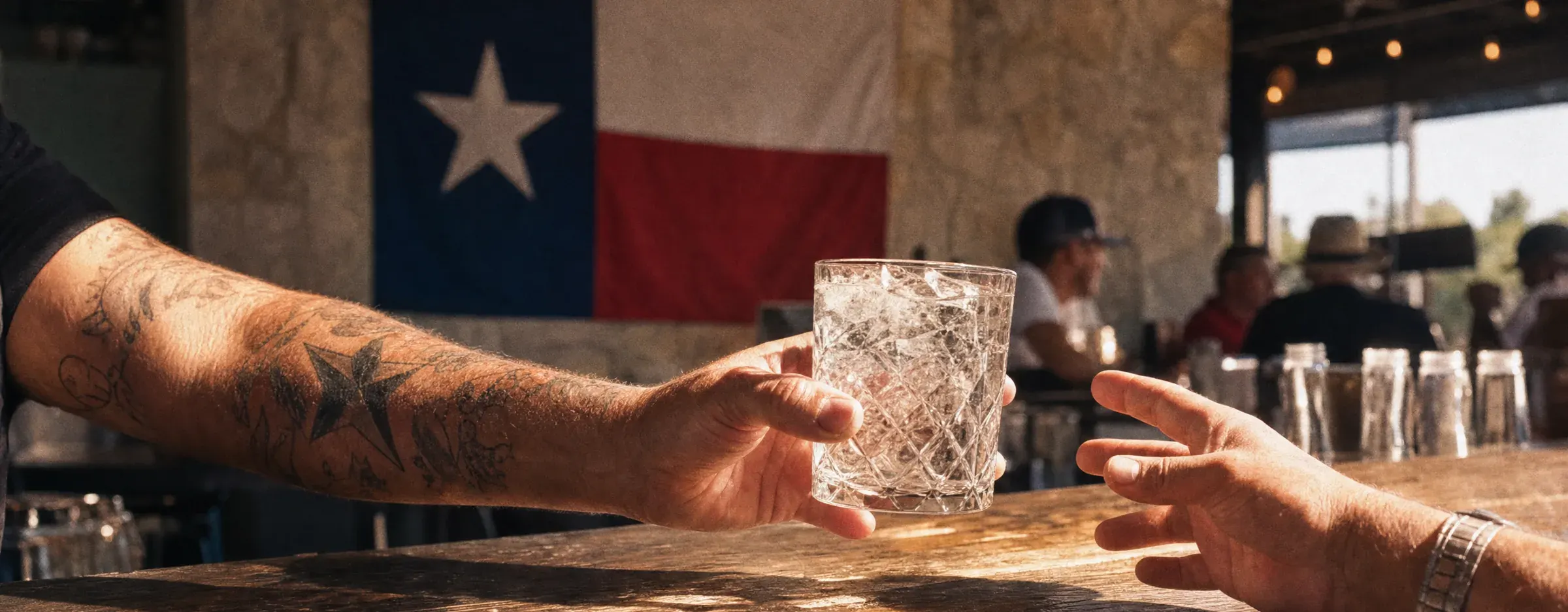 A bartender hands a rocks glass of iced gin across a timber bar under a Texas flag.
