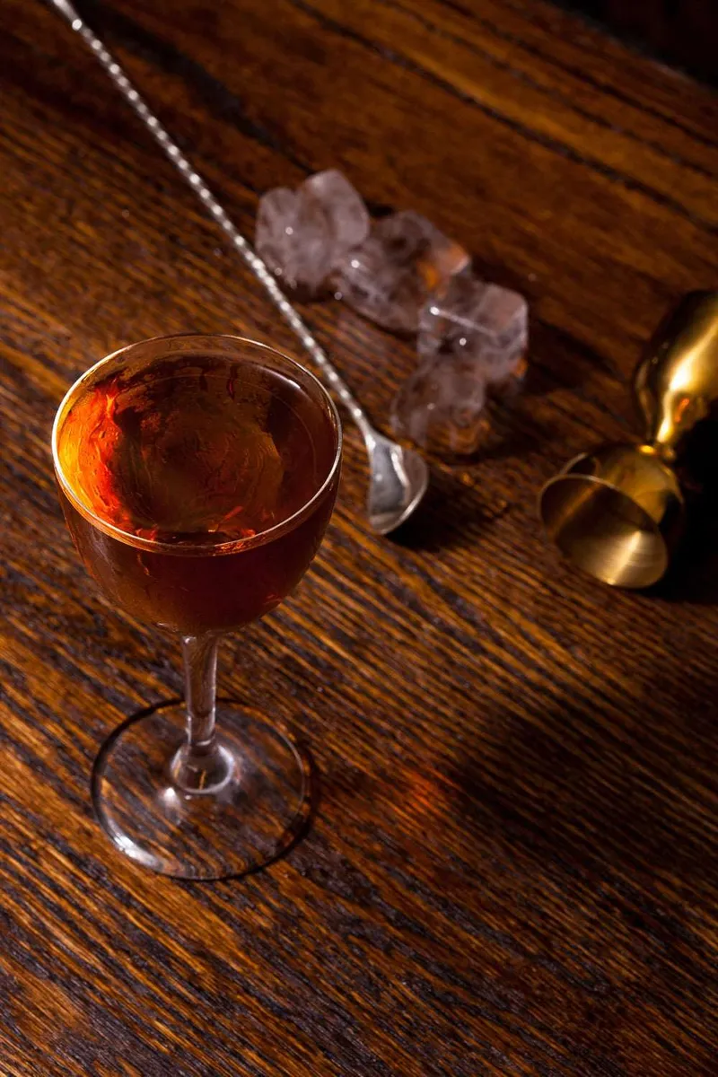 Amber whiskey cocktail in a stemmed glass on a dark wood bar, a bar spoon, ice, and a brass jigger beside it.