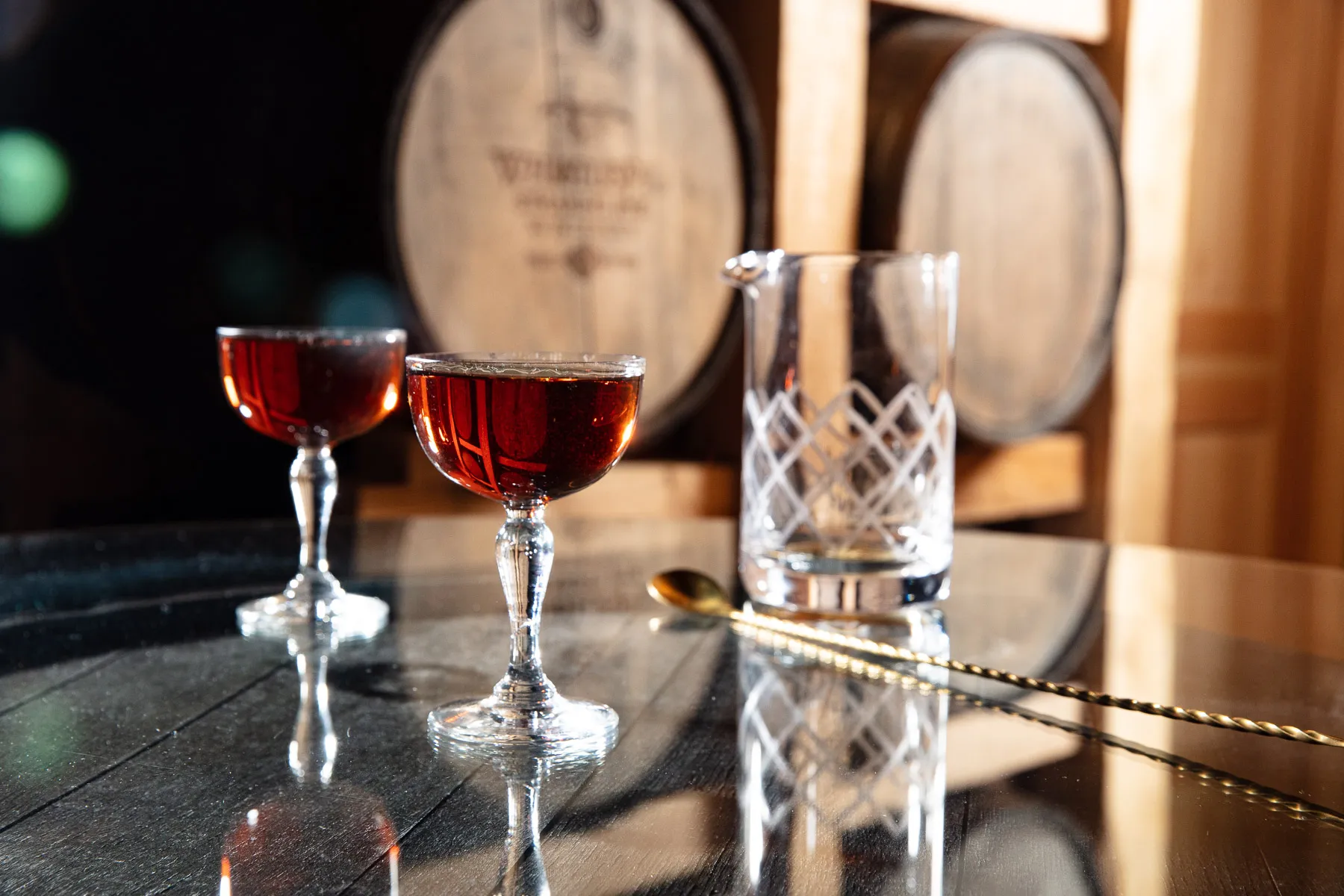 Two stirred cocktails in etched coupes beside a crystal mixing glass on a reflective bar, WhistlePig barrels stacked behind.